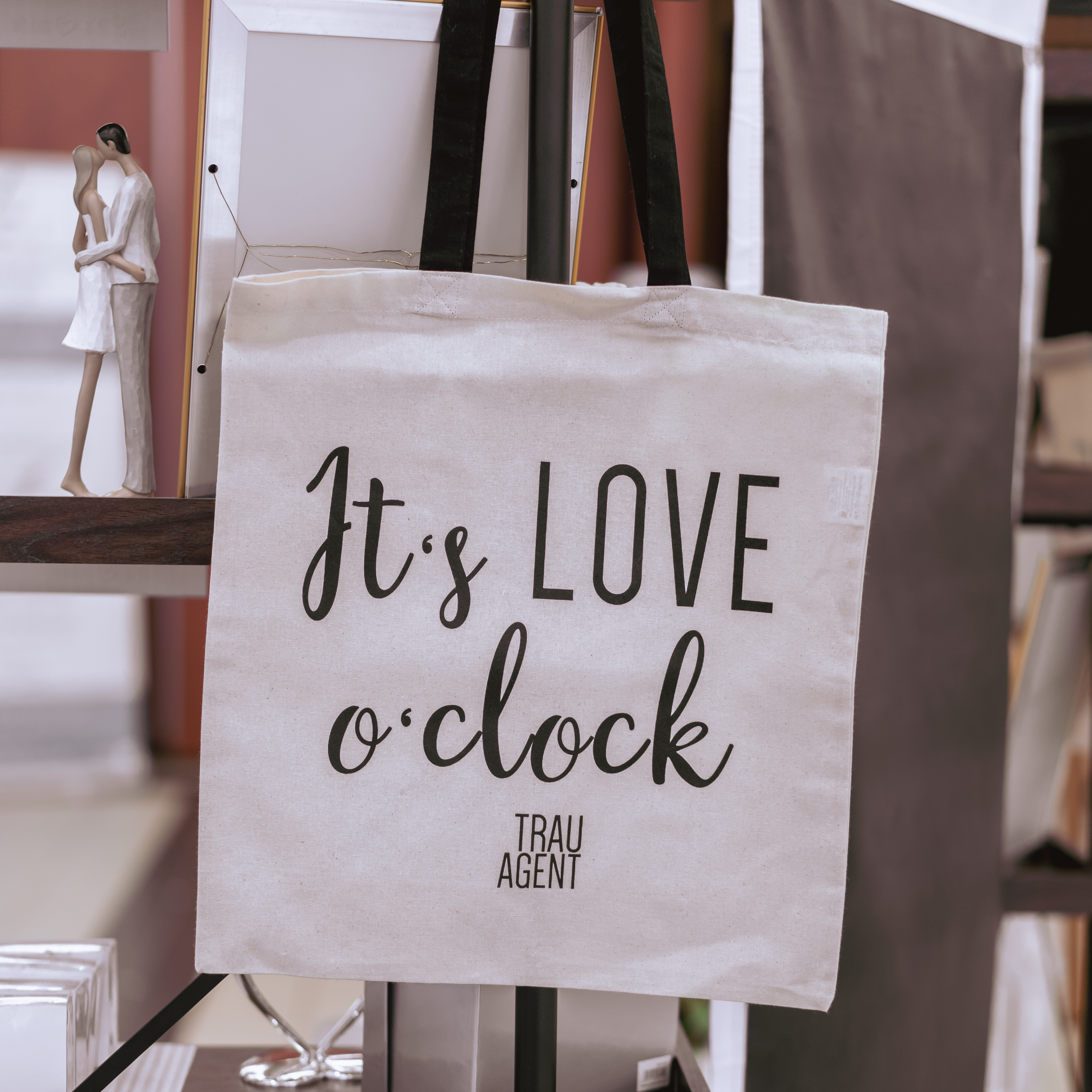 Baumwollbeutel Tasche Aufschrift It's LOVE o'clock mit Trauagent Logo klein aufgehangen  an Dekowand